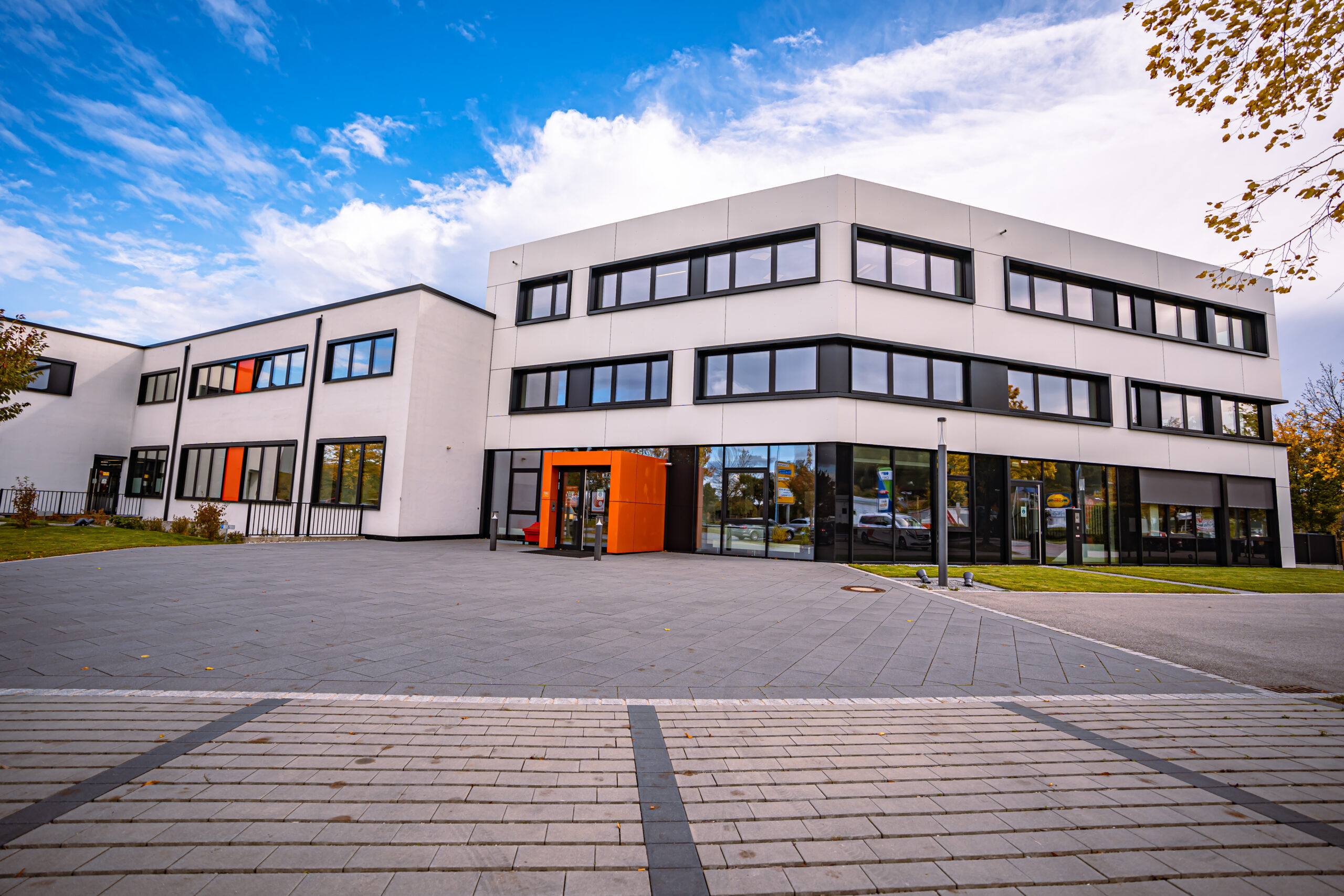 Moderne Unternehmenszentrale mit weißer Fassade, großen Fenstern und markantem orangefarbenem Eingang bei blauem Himmel.