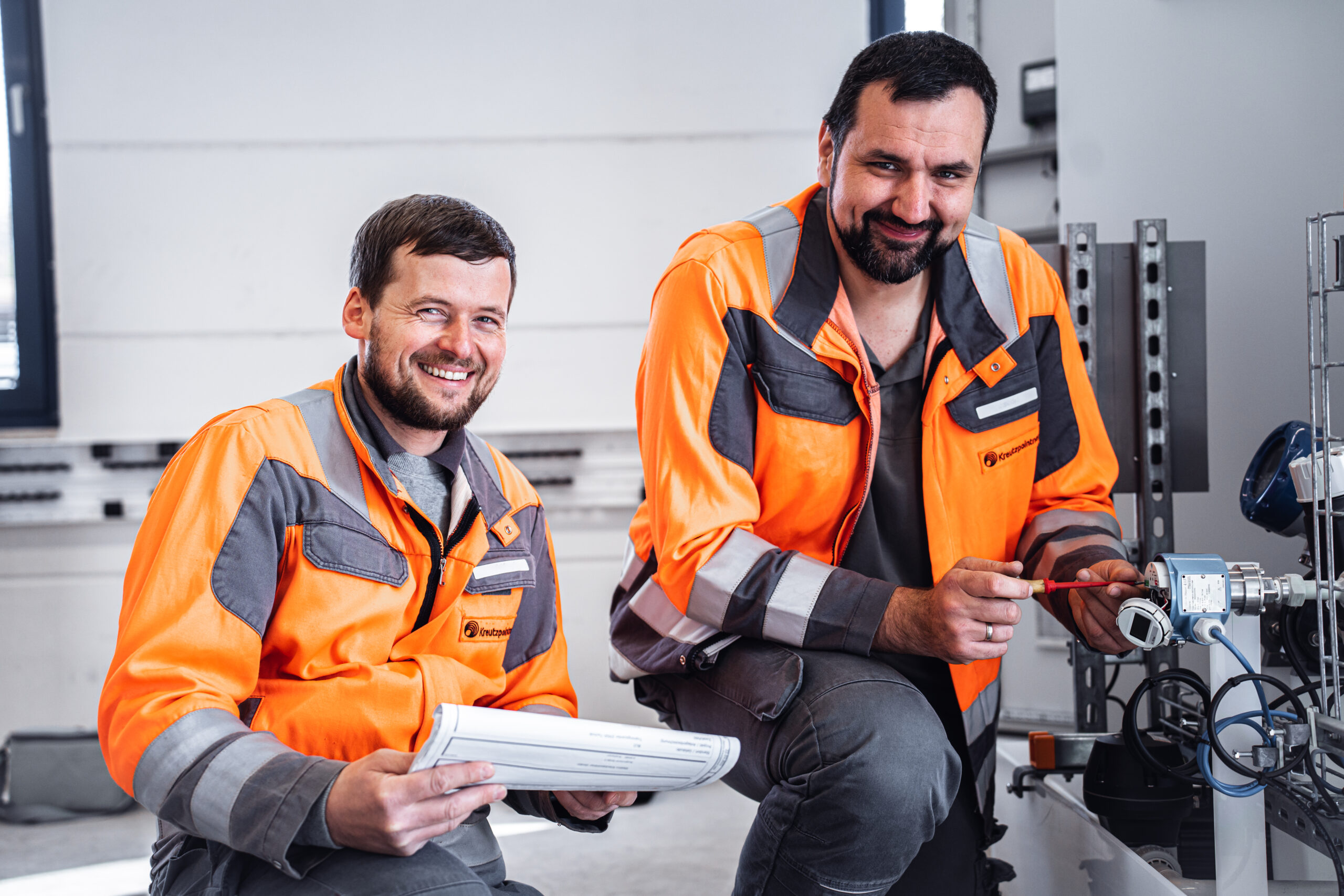 Zwei Männer in orange-grauer Arbeitskleidung knien lächelnd vor einer technischen Anlage, einer hält Baupläne, der andere arbeitet mit Werkzeug am Gerät.