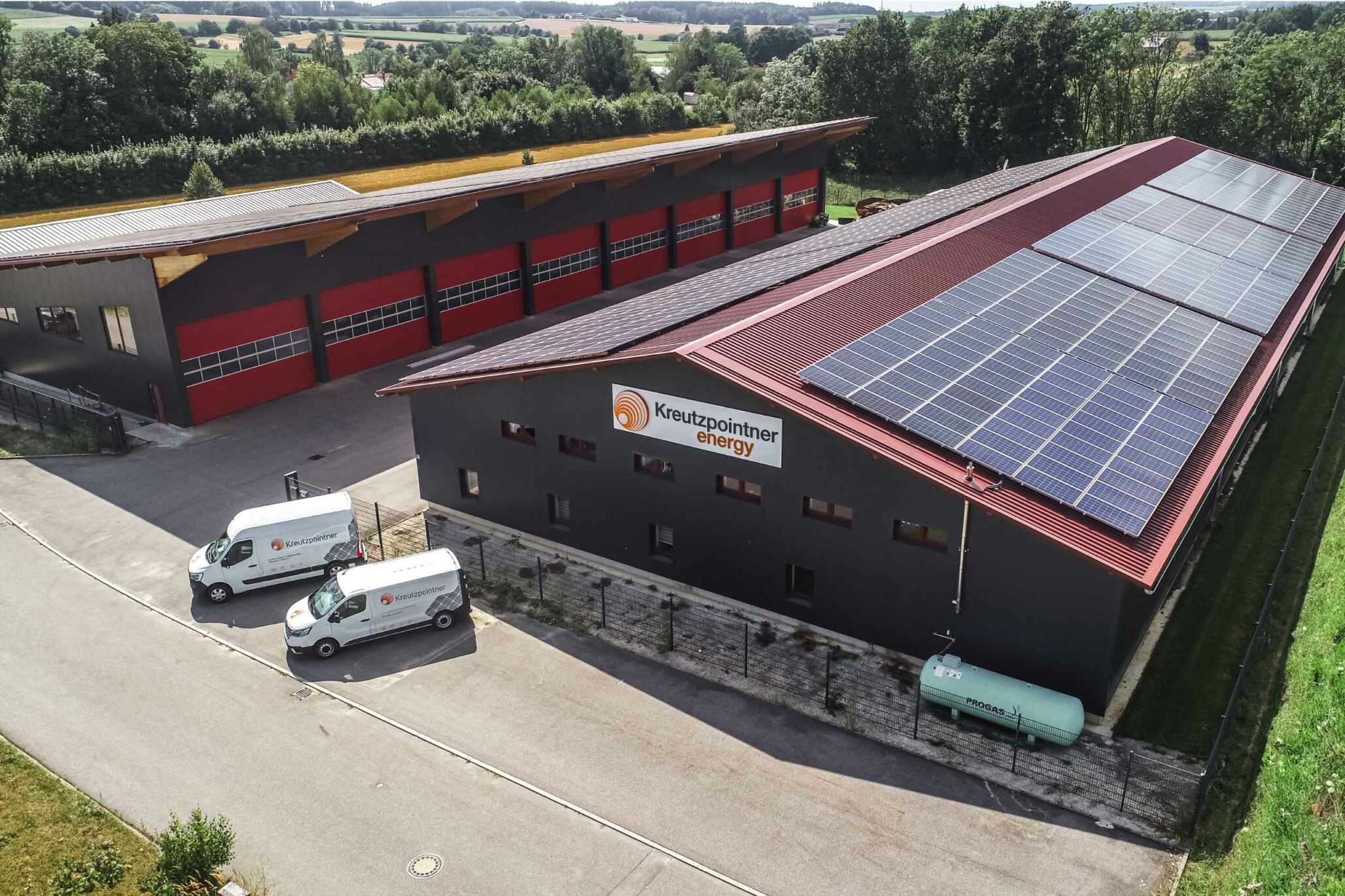 Industriegebäude mit Photovoltaikanlage auf dem Dach, daneben eine große Halle mit roten Rolltoren. Zwei weiße Transporter stehen auf der Straße vor den Gebäuden, im Hintergrund ist eine grüne Landschaft sichtbar.