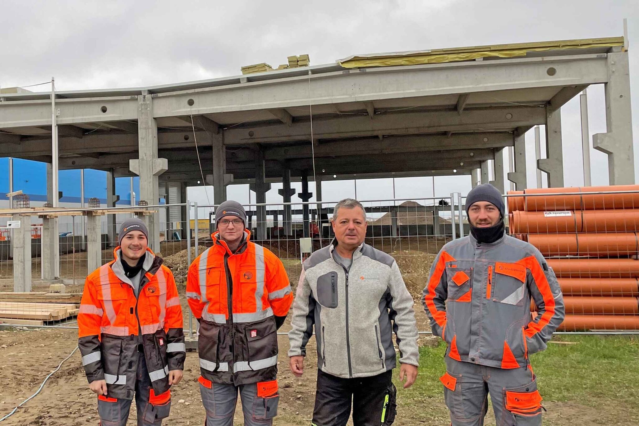 Vier Bauarbeiter in wetterfester Arbeitskleidung stehen vor einem Rohbau mit Betonstützen und offenem Dachtragwerk auf einer Baustelle.