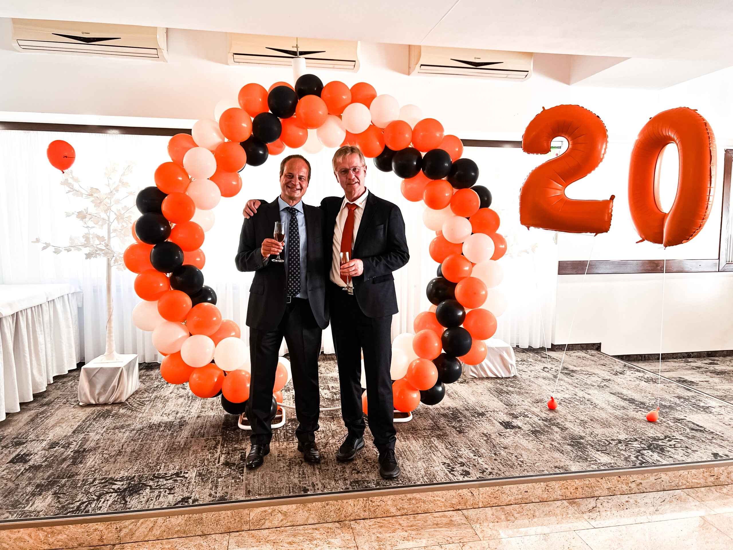 Zwei Männer im Anzug stehen vor einer Ballon-Dekoration in den Farben Orange und Schwarz mit der Zahl „20“.