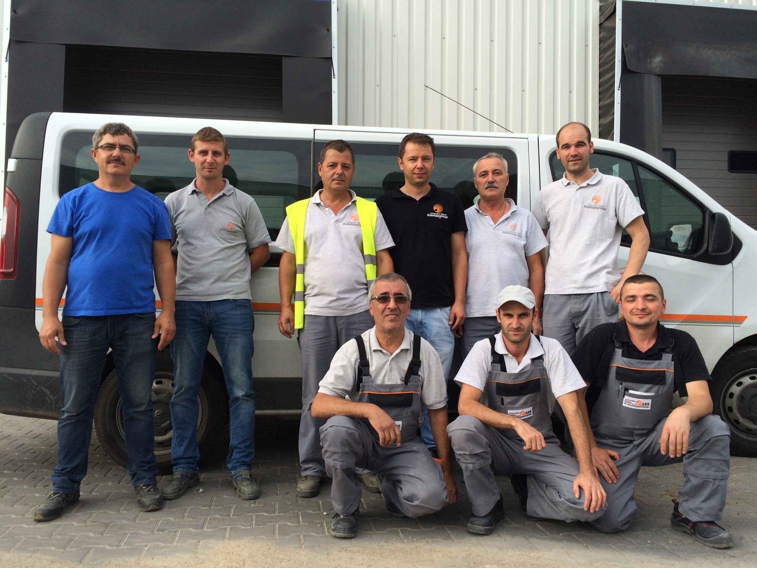 Eine Gruppe von neun Männern in Arbeitskleidung steht und hockt vor einem hellen Industriegebäude für ein Gruppenfoto.