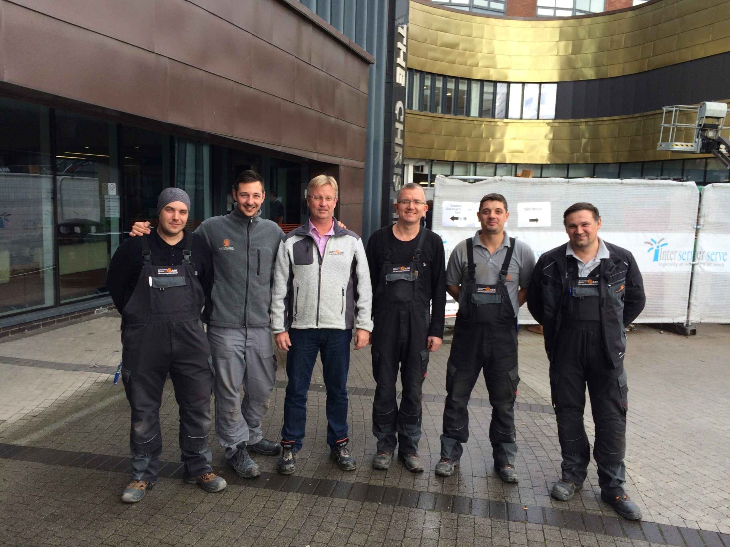 Sieben Mitarbeiter in dunkler Arbeitskleidung posieren für ein Gruppenfoto vor einem industriellen Hintergrund.