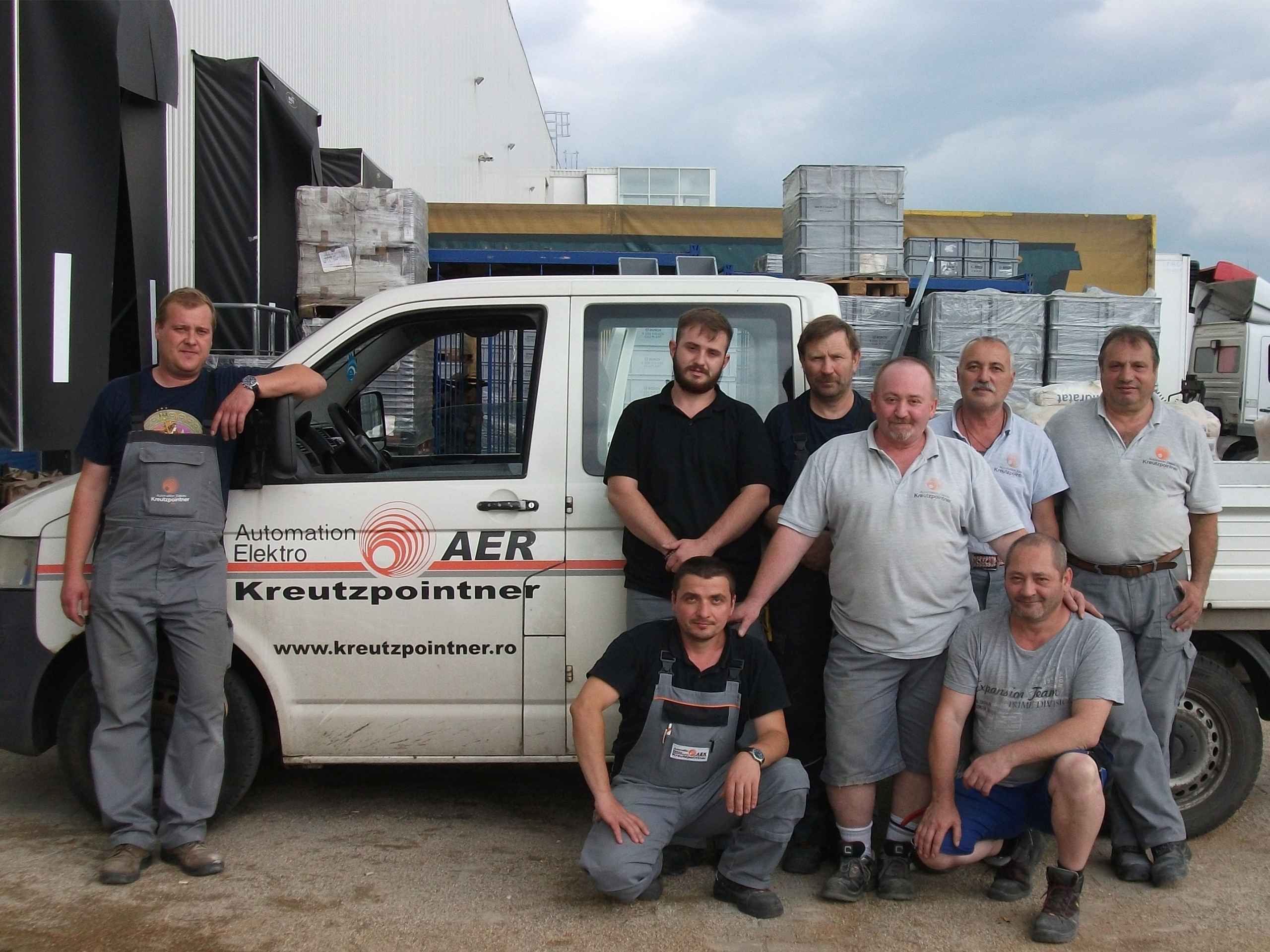 Eine Gruppe von acht Männern steht und hockt neben einem weißen Firmenfahrzeug mit Kreutzpointner-Logo und der Aufschrift „AER“.