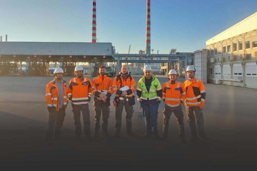 Gruppenfoto von sechs Industriearbeitern in Schutzkleidung und Helmen auf dem Außengelände eines Industriekomplexes mit Schornsteinen im Hintergrund.