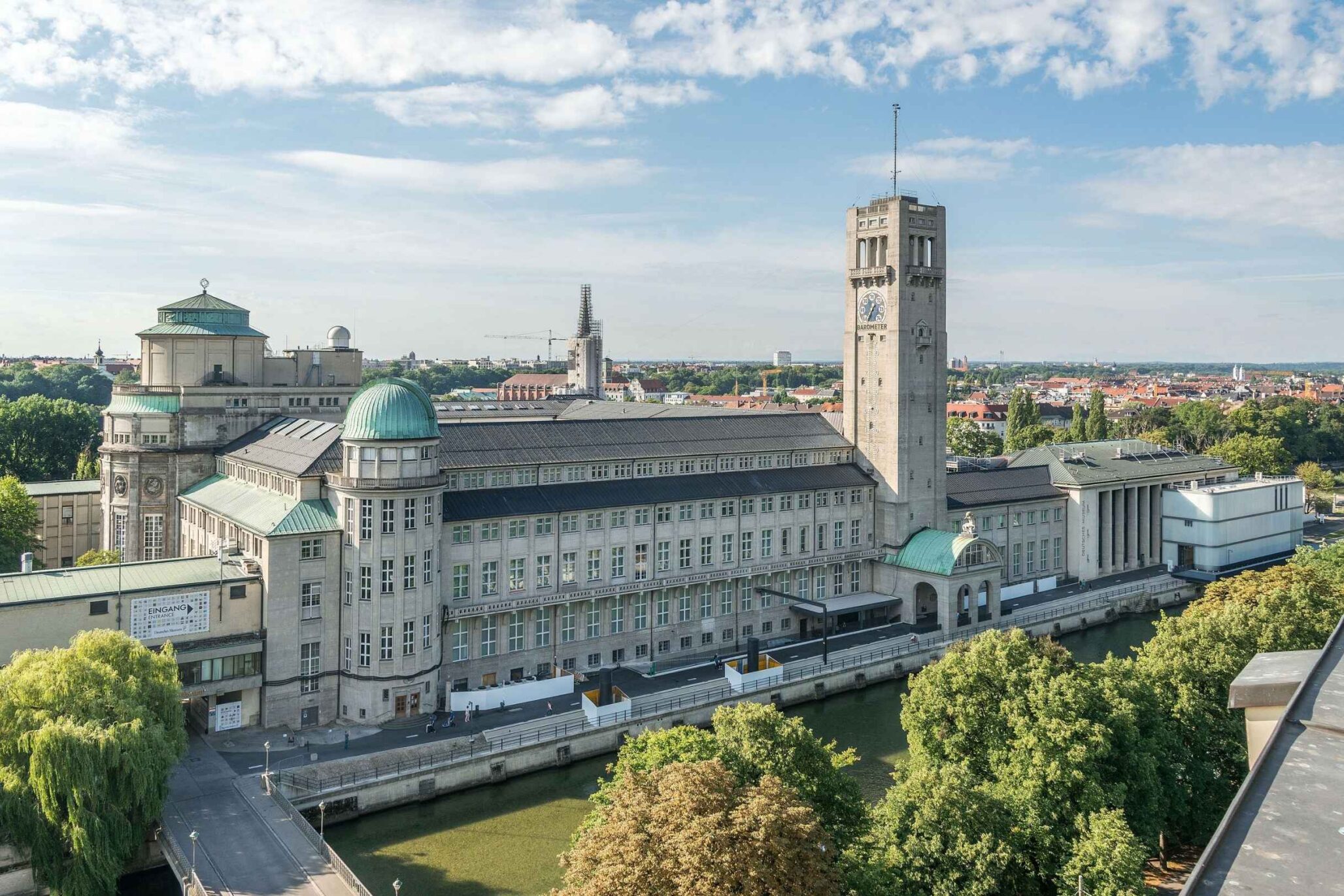 Außenansicht des Deutschen Museums in München mit Isar im Vordergrund und Blick auf die Stadt bei sonnigem Wetter.