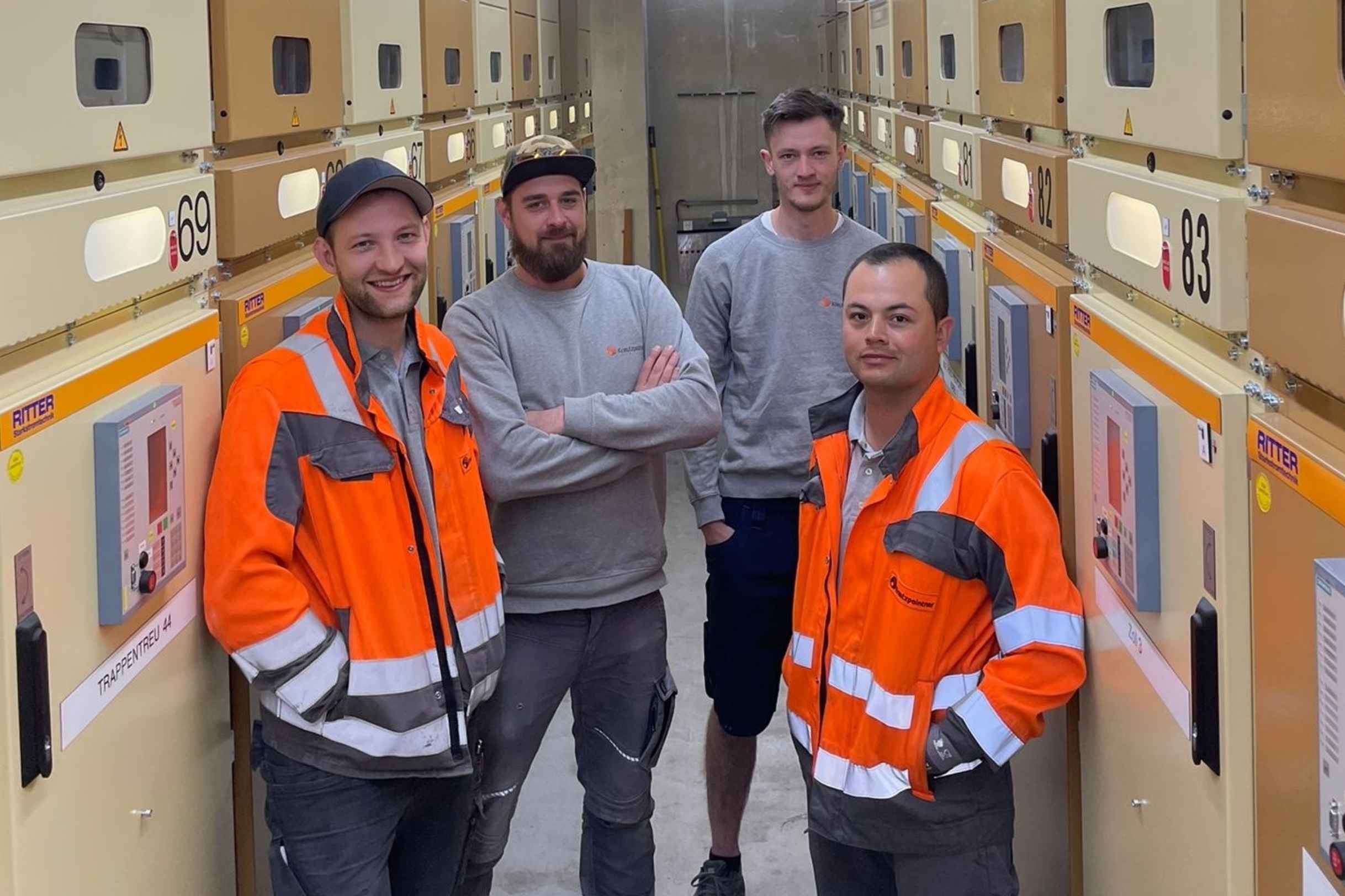 Vier Elektriker stehen in einem Technikraum zwischen Schaltanlagen; zwei tragen orange Warnschutzjacken, zwei in grauer Arbeitskleidung.