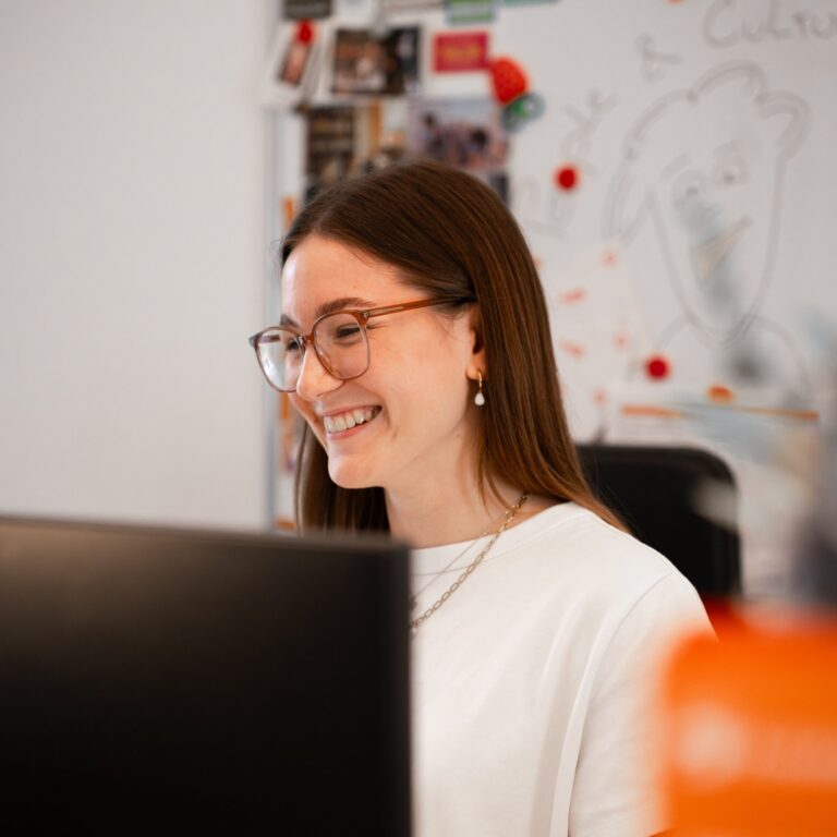 Junge Frau mit Brille und langen braunen Haaren lächelt vor einem Bildschirm in einem Büro; im Hintergrund ist ein Whiteboard mit Zeichnungen und Fotos zu sehen.