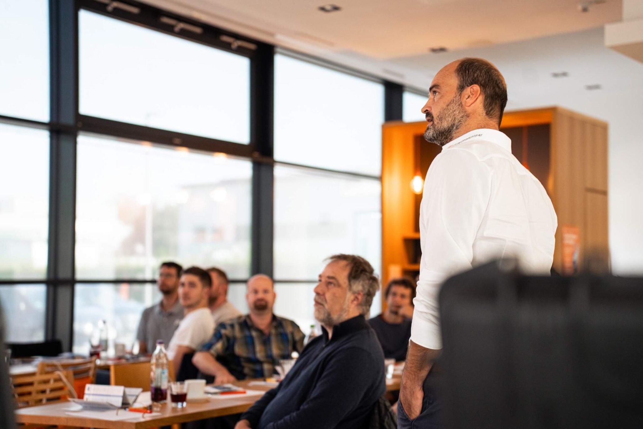 Ein Dozent hält eine Präsentation vor einer aufmerksam zuhörenden Gruppe in einem modernen, hellen Seminarraum mit großen Fensterfronten.