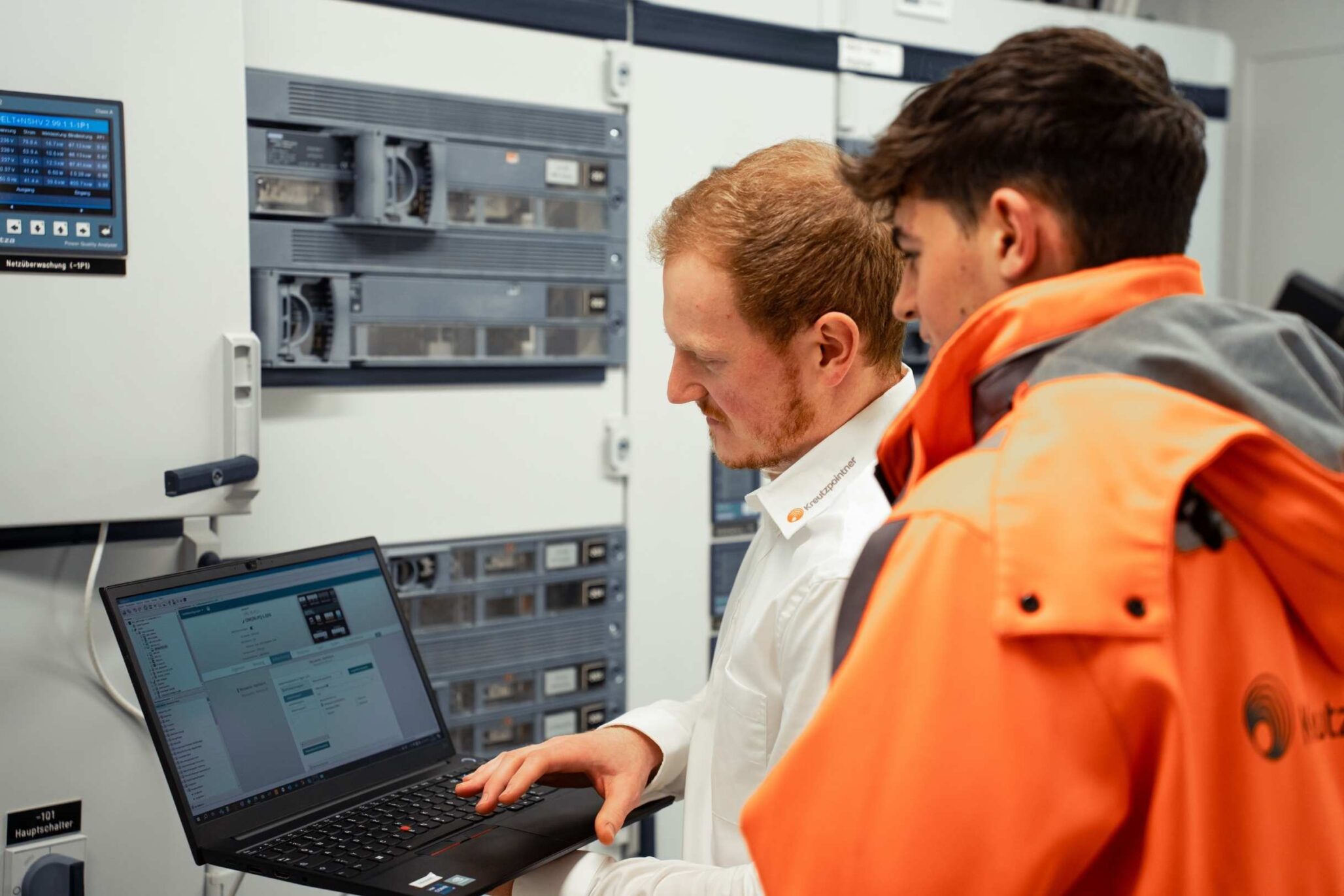 Zwei Techniker prüfen an einem Laptop die Konfiguration einer Schaltanlage im Schaltraum; einer trägt orangefarbene Arbeitskleidung.
