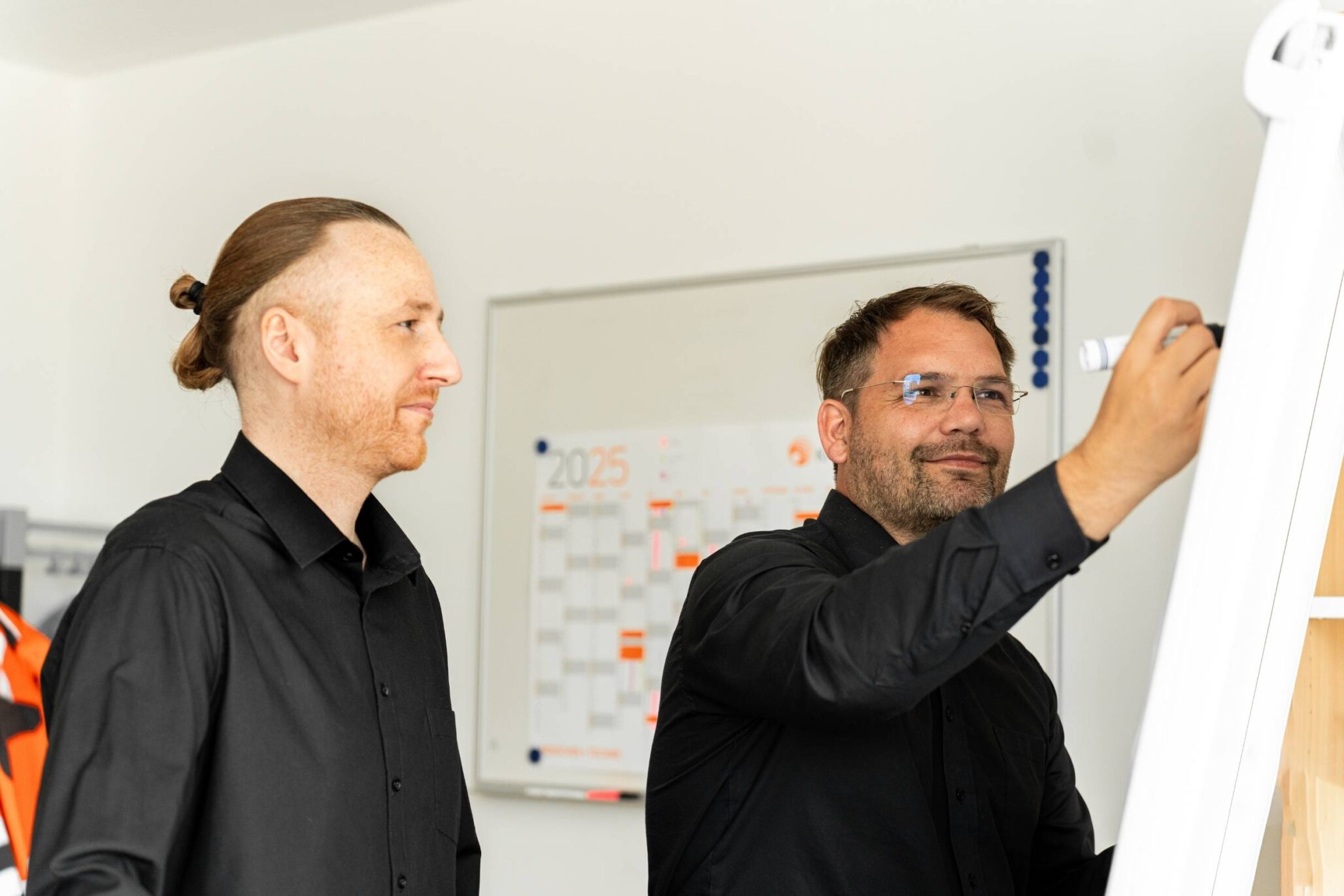 Zwei Männer in schwarzer Kleidung arbeiten gemeinsam an einem Flipchart in einem Büro mit Wandkalender im Hintergrund.