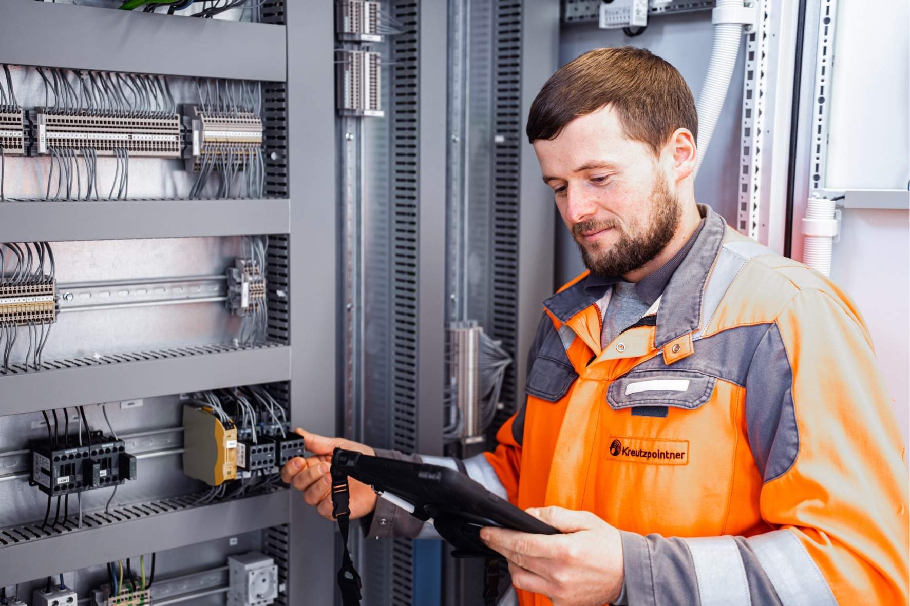 Techniker in orange-grauer Arbeitskleidung mit einem Tablet vor einem geöffneten Schaltschrank, prüft elektrische Verbindungen und Komponenten.