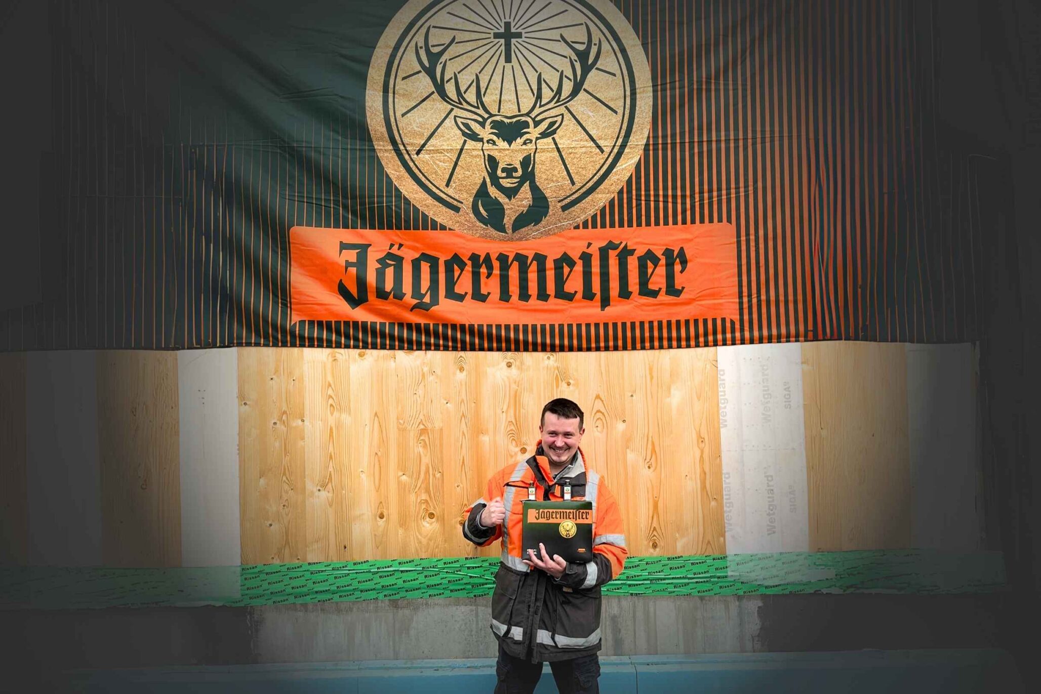 Mann in orangefarbener Arbeitskleidung steht vor einem großen Jägermeister-Banner und hält eine Geschenkbox der Marke in der Hand.