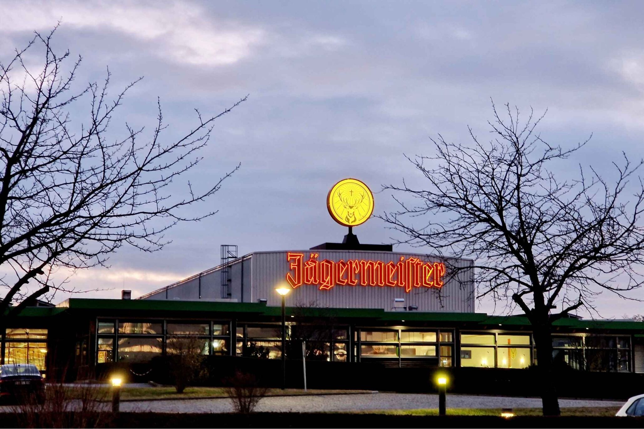 Außenansicht des Jägermeister-Werks in Kamenz bei Dämmerung, mit beleuchtetem Logo und winterlicher Baumkulisse.