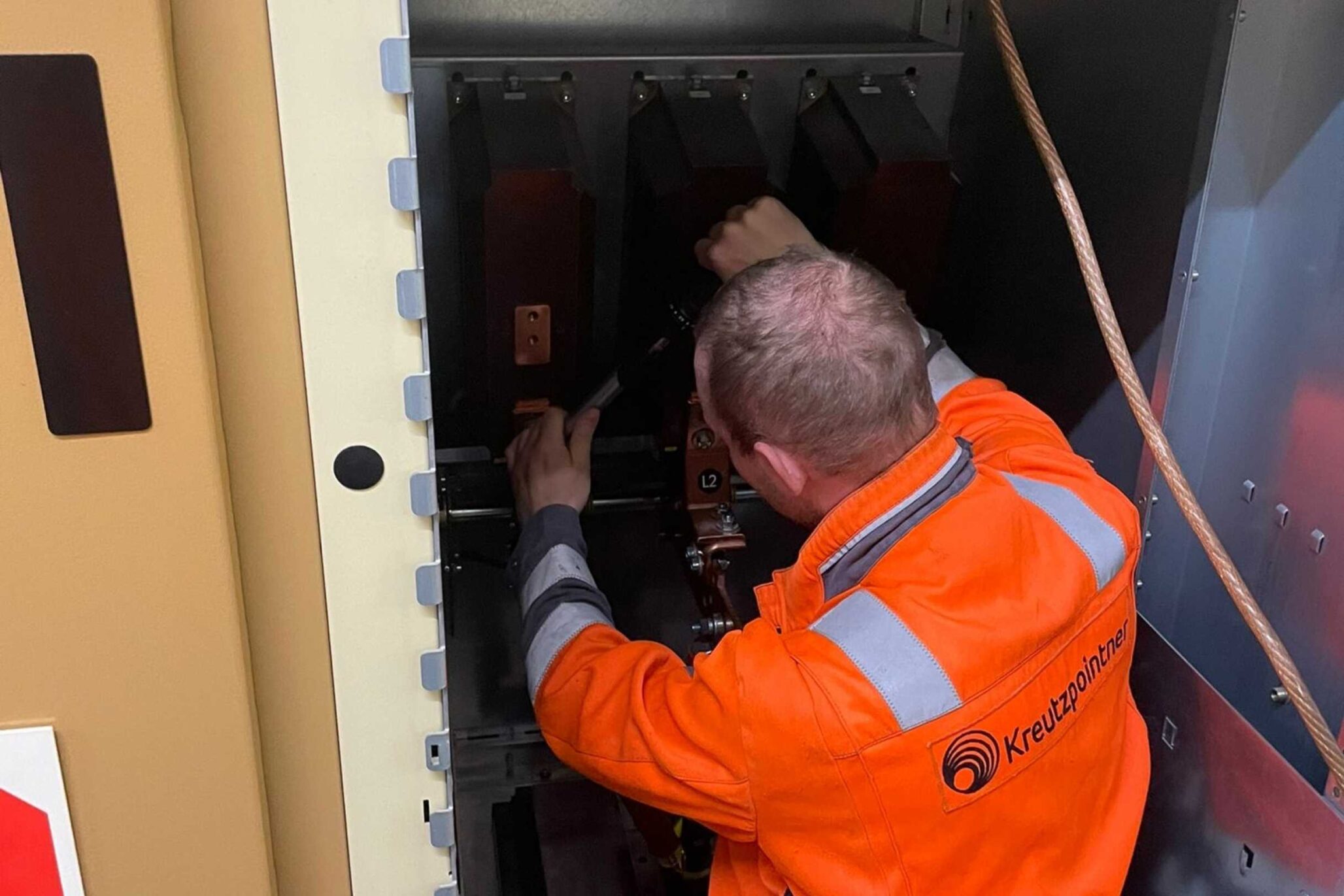 Techniker in orangefarbener Warnschutzkleidung arbeitet im Inneren einer geöffneten Mittelspannungsschaltanlage an elektrischen Komponenten.