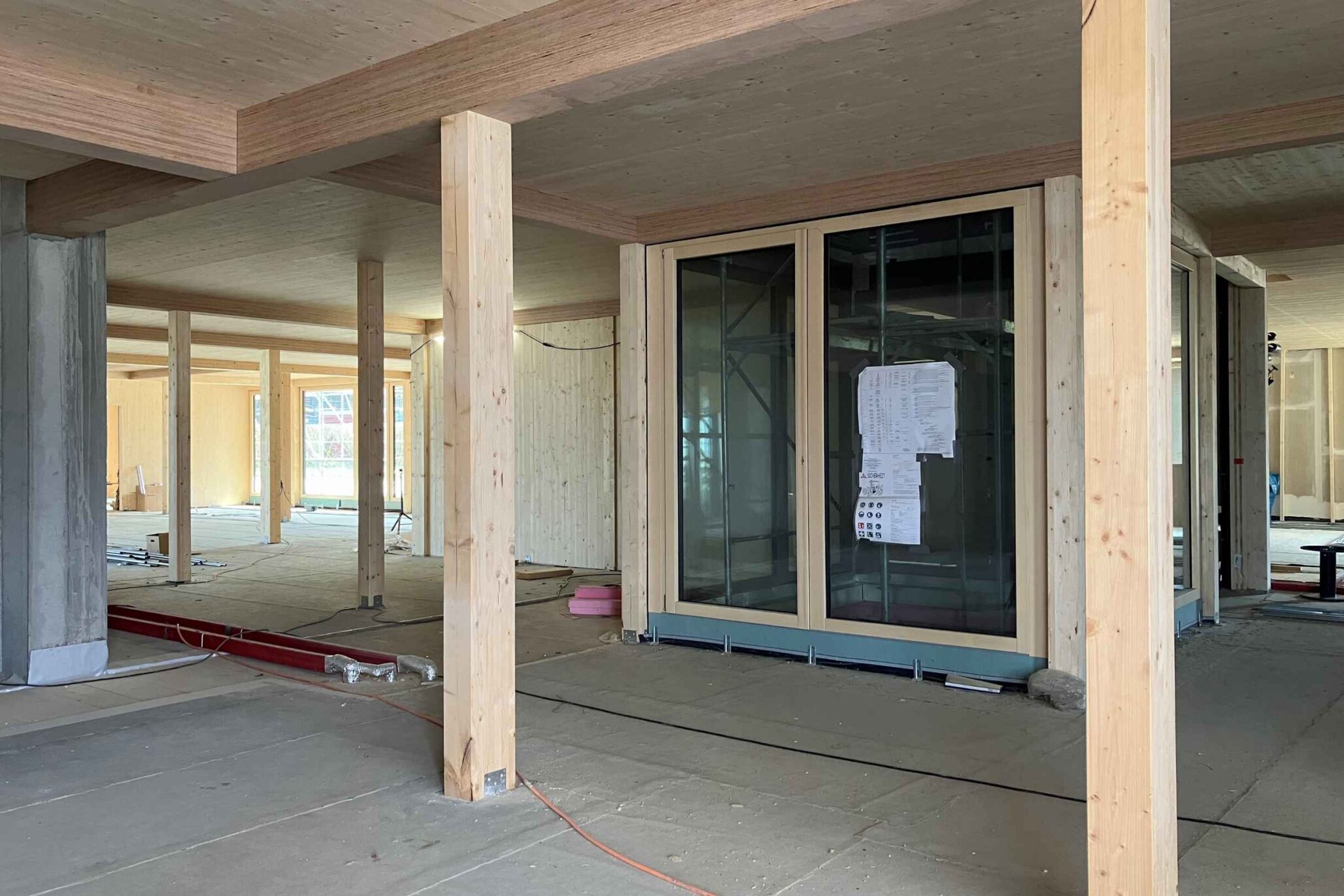 Innenaufnahme eines Gebäuderohbaus mit Holzstützen und vorbereiteter Installationsebene, Fensterfront im Hintergrund.