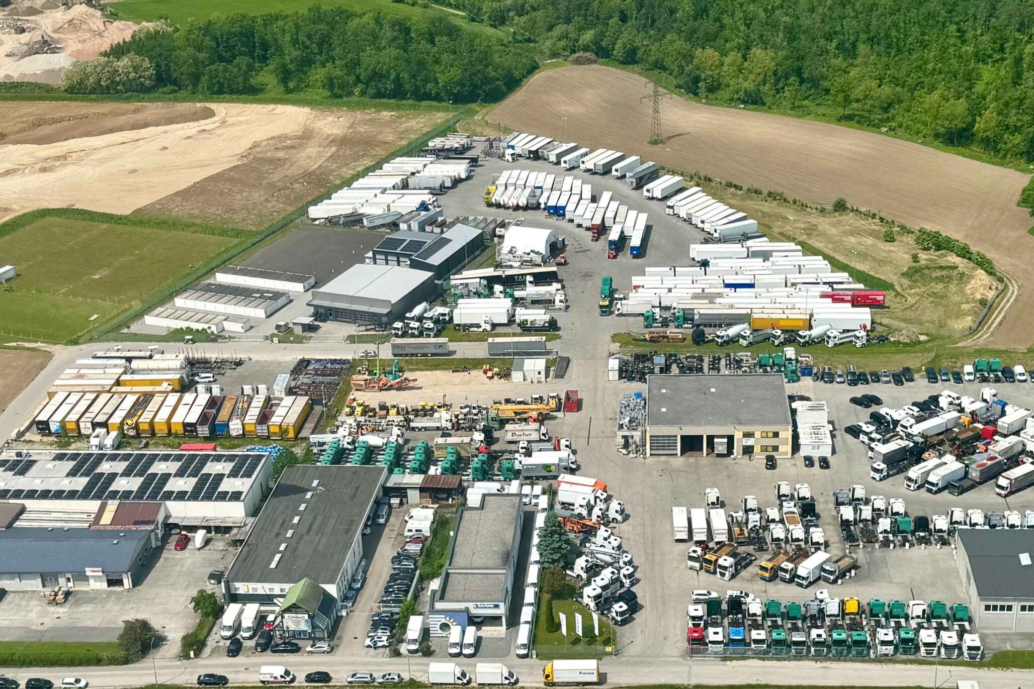 Luftaufnahme eines weitläufigen Gewerbe- und Logistikgeländes mit zahlreichen Lkw, Anhängern, Lagerhallen und Freiflächen, umgeben von Feldern und Wald.