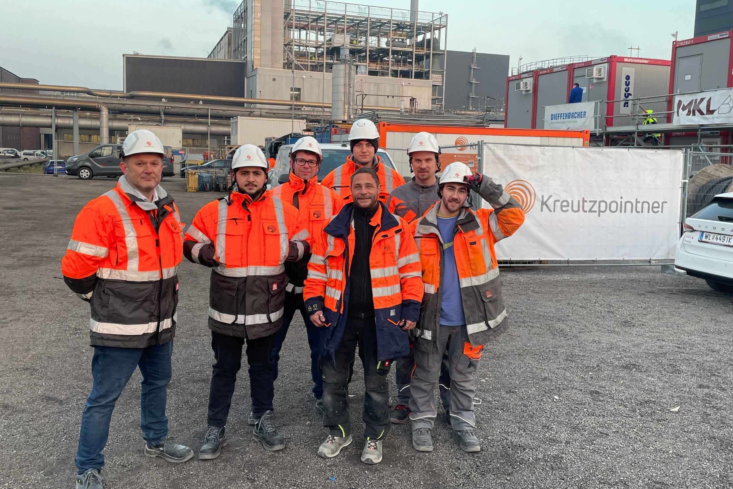 Gruppenfoto von sieben Bauarbeitern in orangefarbener Schutzkleidung und Helmen auf einer Baustelle der Firma Elektro Kreutzpointner Austria GmbH, mit Industrieanlagen im Hintergrund.