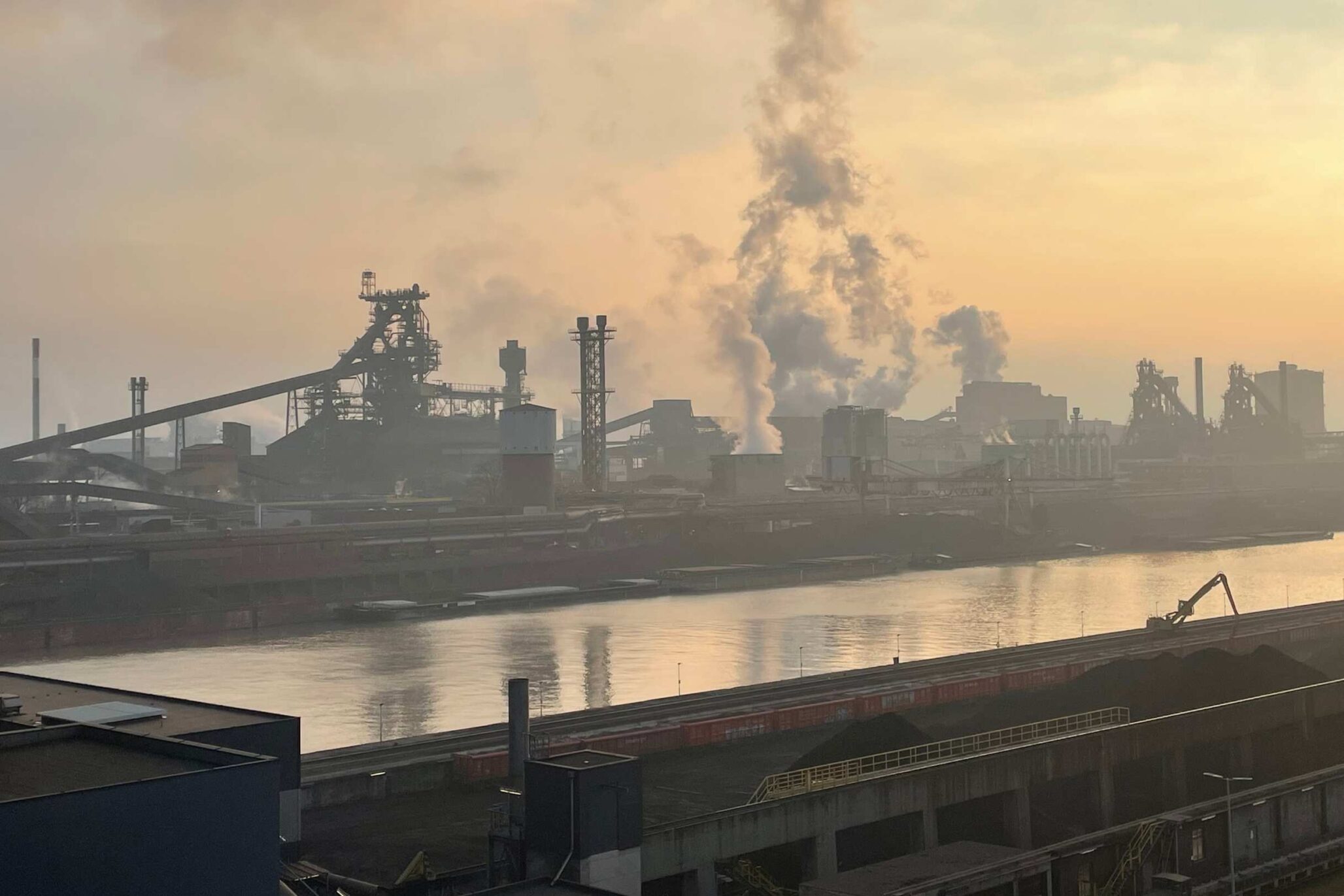Industriegelände mit rauchenden Schornsteinen an einem Flussufer bei Sonnenaufgang – Blick auf eine Stahl- oder Schwerindustrieanlage mit starkem Ausstoß von Wasserdampf oder Rauch.