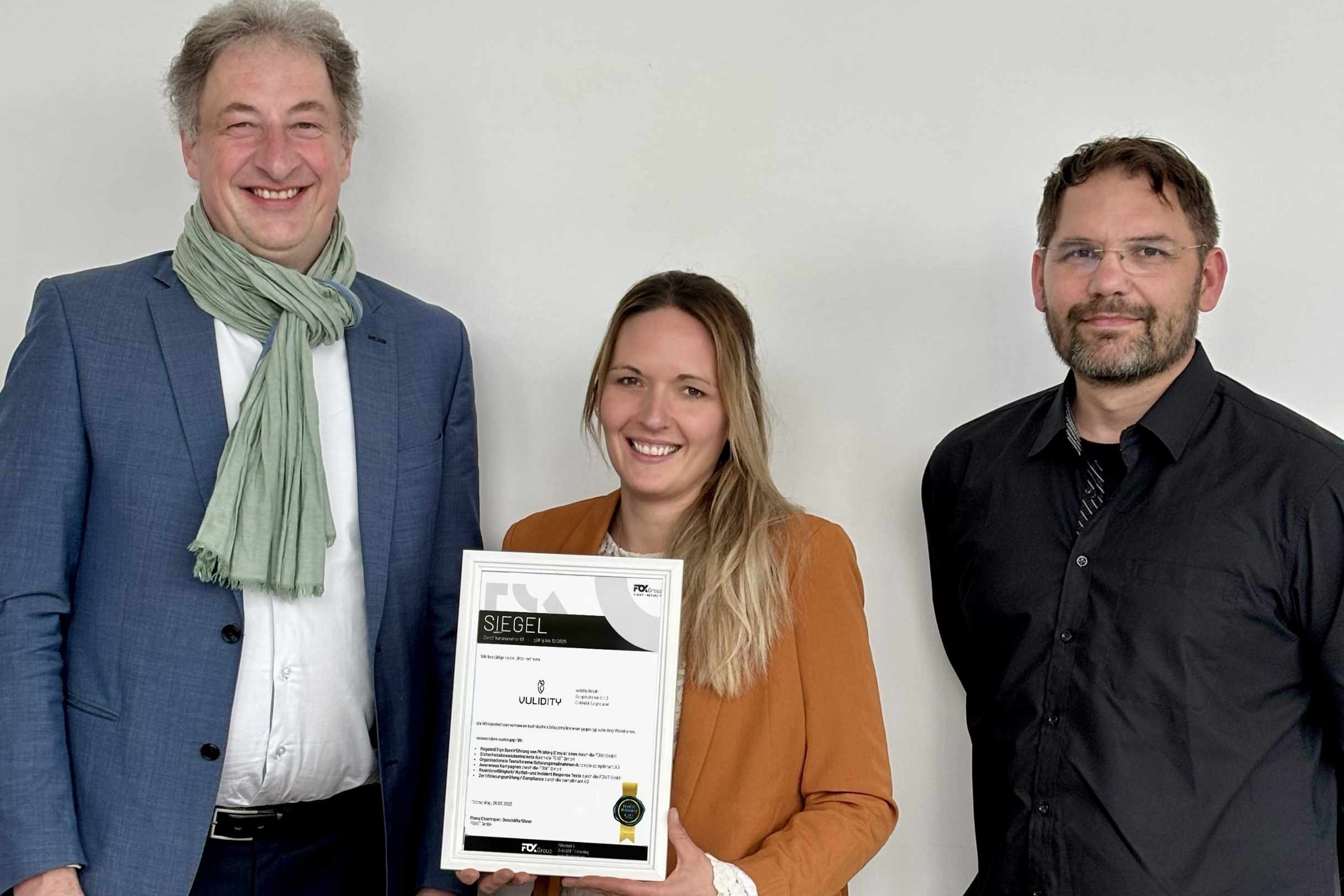 Drei Personen präsentieren stolz das neu entwickelte Vulidity-Siegel, eine von ihnen hält eine gerahmte Urkunde mit Zertifikat in der Hand.
