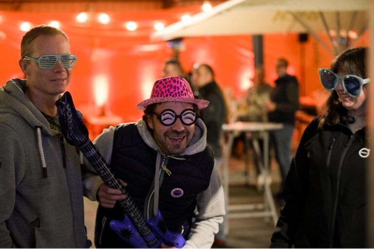 Bei abendlicher Stimmung spielt ein Herr neben einem weiteren Herren und eine Frau mit lustigen Hüten und Brillen eine Luftgitarre