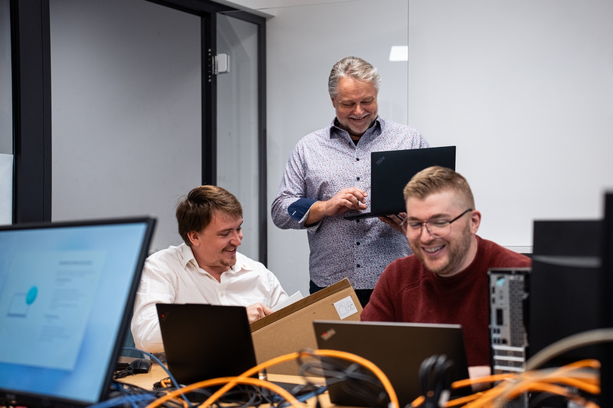 Drei Männer sitzen in einem IT Testraum und arbeiten an Laptops.