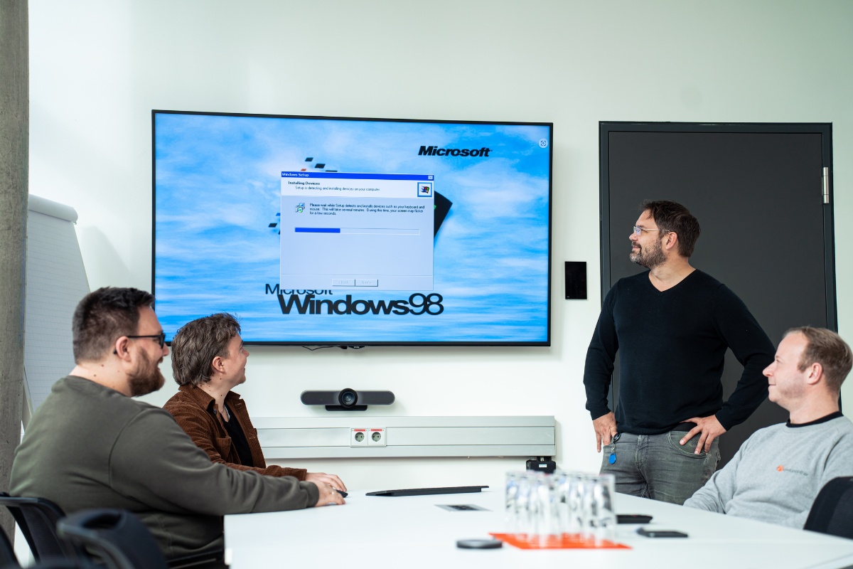 4 Männer, davon einer stehend und 3 sitzend schauen auf einen Bildschirm auf dem gerade ein Windows Update läuft.