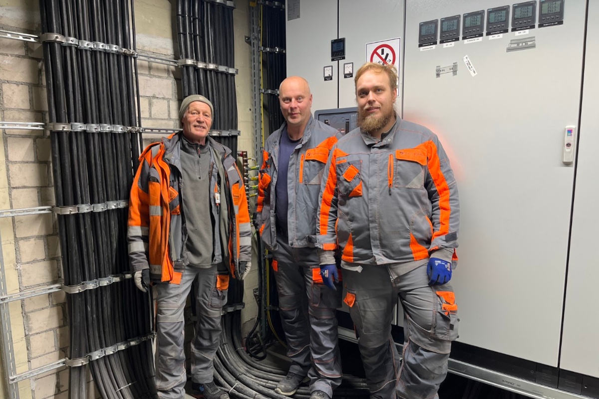 3 Männer in Arbeitskleidung stehen vor einen Schaltschrank und zwei Kabeltrassen an der Wand.