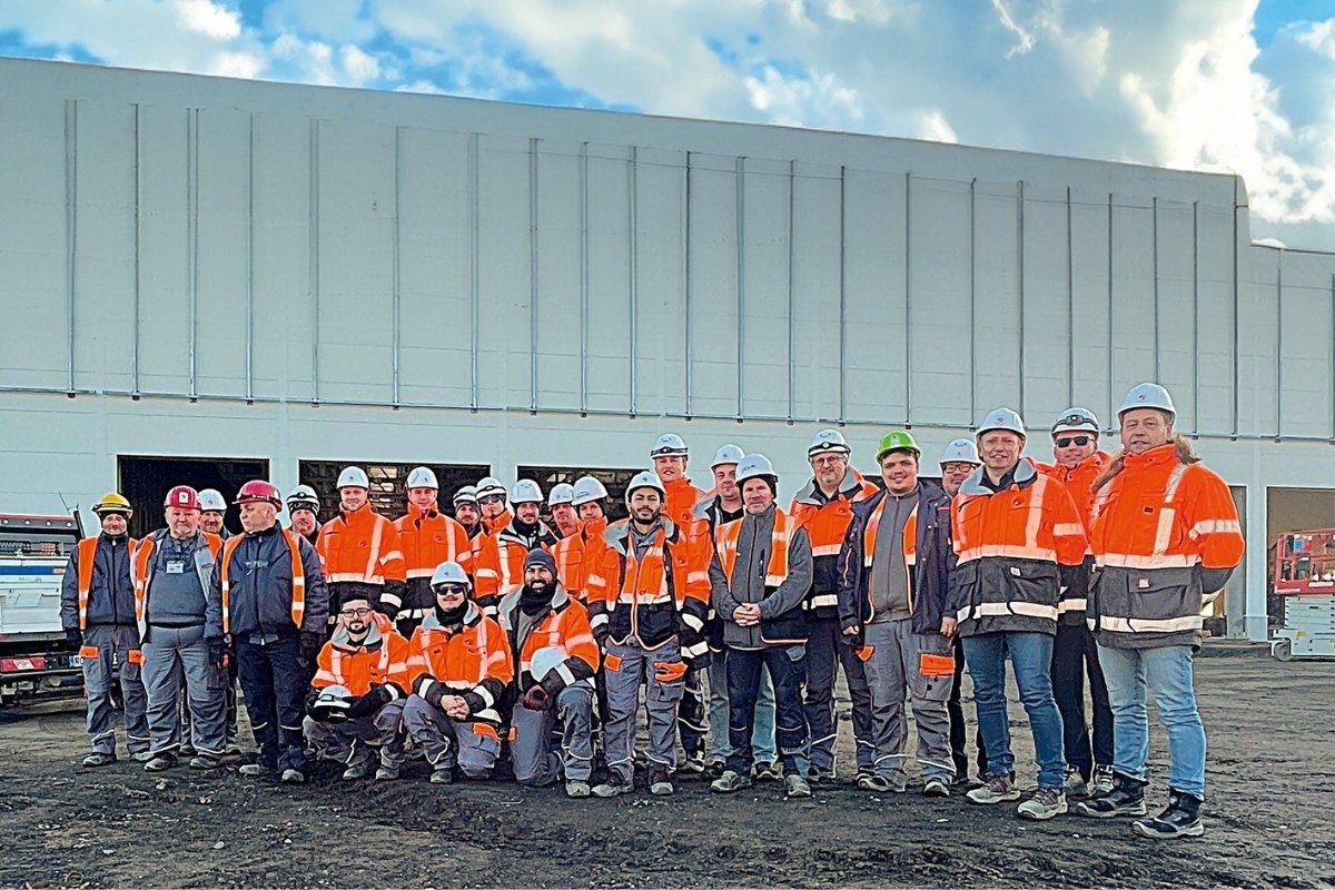 Mannschaftsfoto in Arbeitskleidung vor einer großen Halle mit sonnigem Himmel und Wolken.