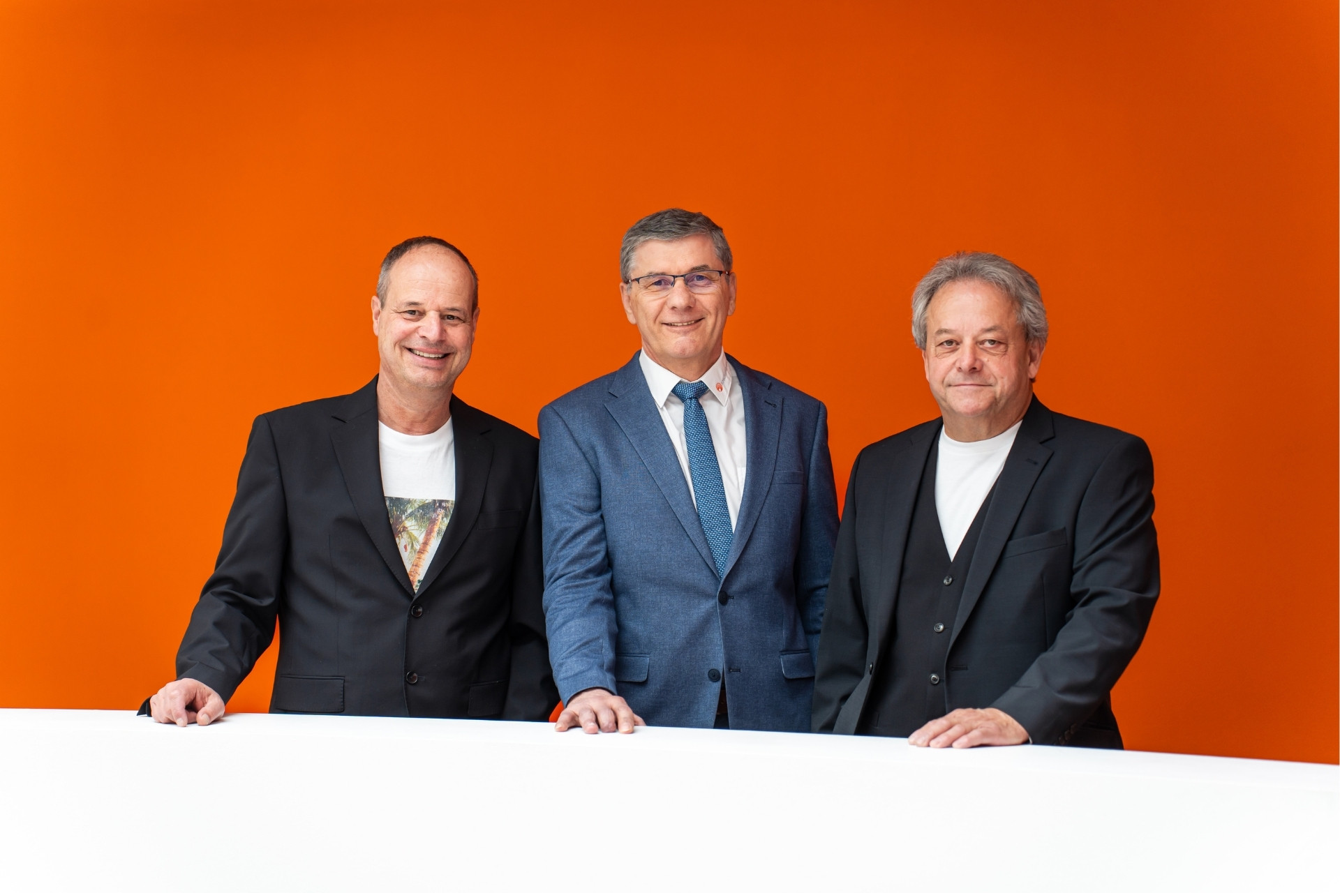Drei Herren im Azubi stehen vor einer orangenen Wand und lachen in die Kamera.