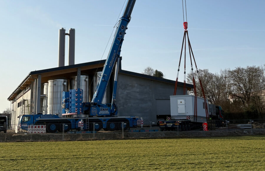 Ein Krahn hebt einen Trafo vor einer großen Halle, die sich aber noch im Bau befindet.