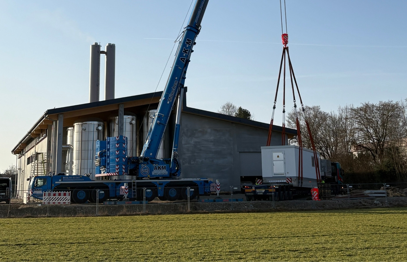 Ein Krahn hebt einen Trafo vor einer großen Halle, die sich aber noch im Bau befindet.