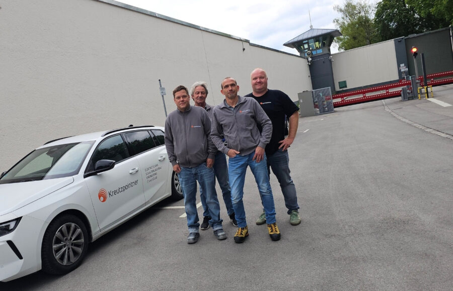 Vier Männer stehen vor einer weißen Mauer, die zu einem Gefängnis gehört. Sie stehen neben einem Firmenfahrzeug für ein Gruppenfoto.
