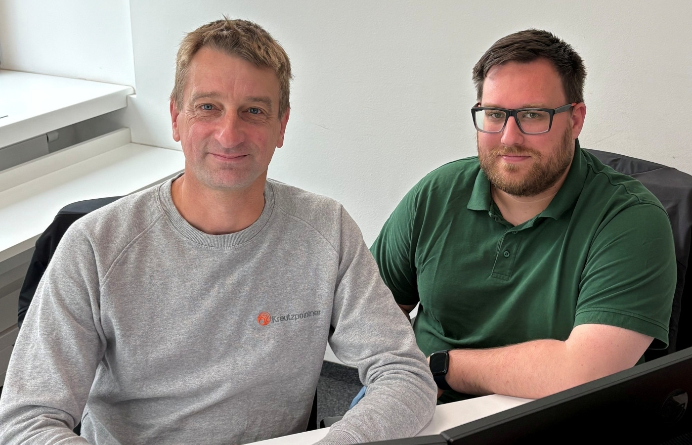 Teamfoto von zwei Herren die am Schreibtisch vor einem PC sitzen aber in die Kamera schauen.