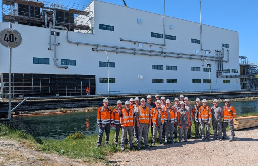 Gruppenfoto von Männern in Arbeitskleidung vor einem Werk, wo davor noch Wasser fließt.