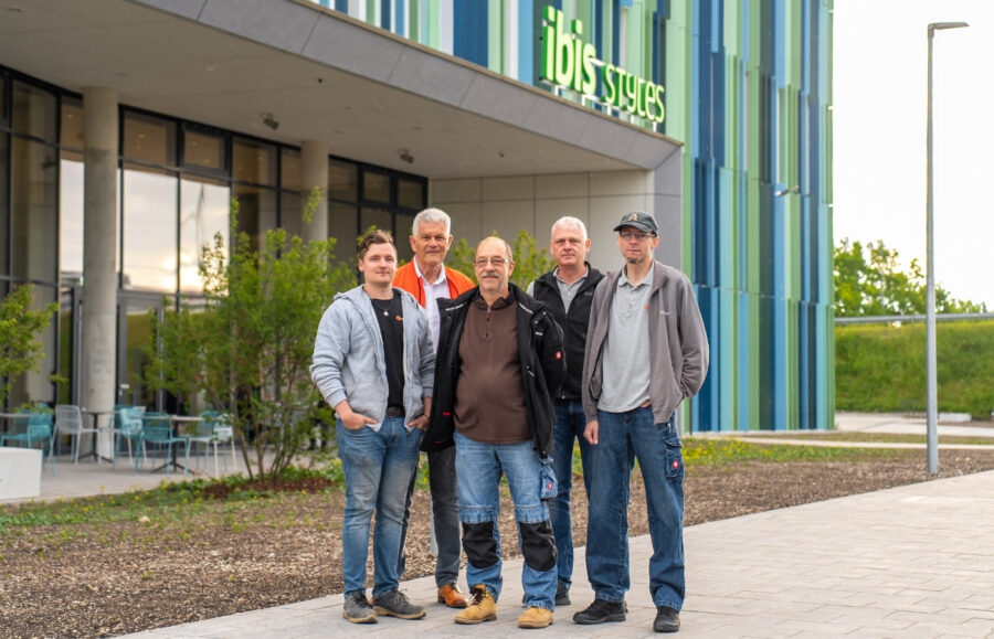 Fünf Herren stehen für ein Gruppenbild vor einem Ibis Hotel, das eine blau-grüne Fassade hat.
