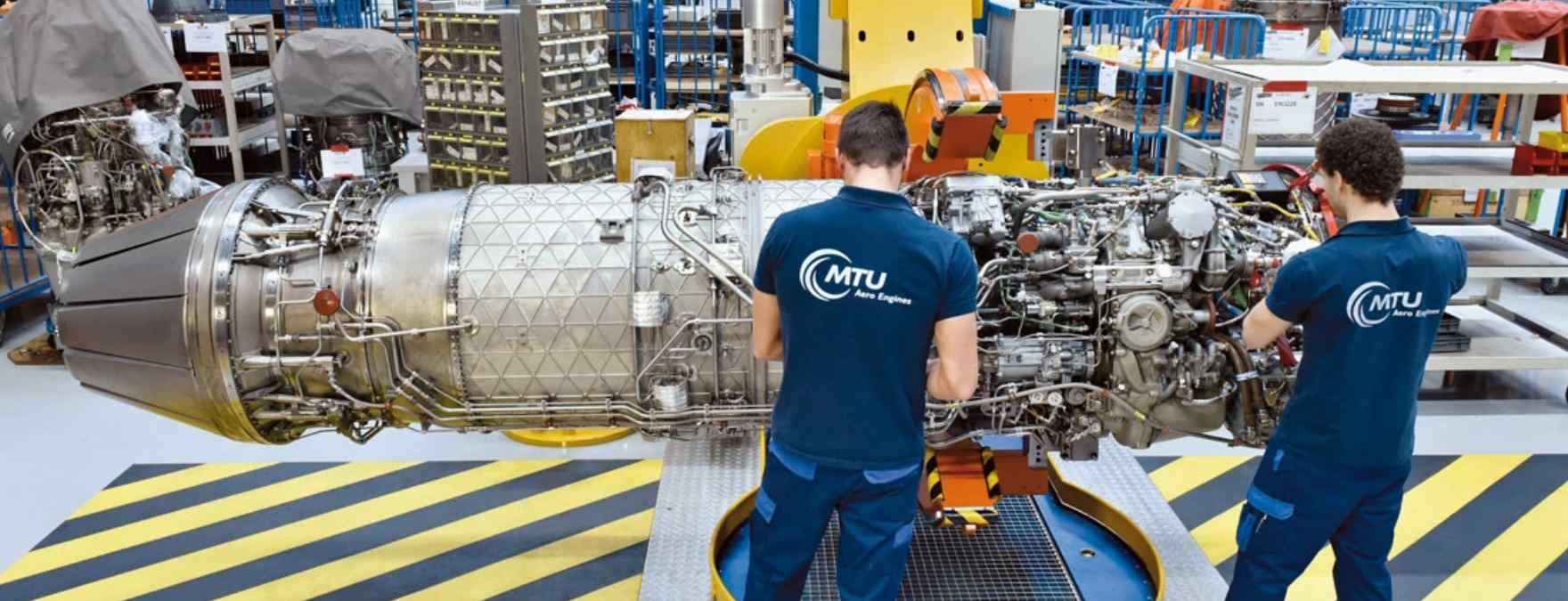 Zwei Männer in Arbeitskleidung stehen vor einem riesen Triebwerk und arbeiten daran.
