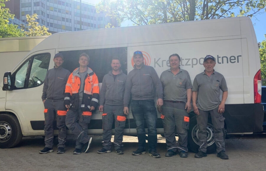 Sechs Männer in Arbeitskleidung stehen vor einem Baustellenfahrzeug der Firma Kreutzpointnter. Es scheint die Sonne und im Hintergrund sieht man ein großes Gebäude.