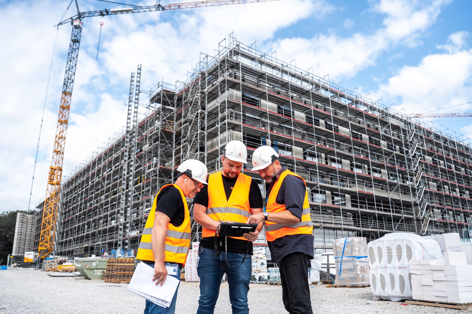 Drei Herren in Arbeitskleidung stehen vor einer Baustelle. Ein großes Gebäude wird errichtet mit Krahn und Baustoffen ausßenrum.