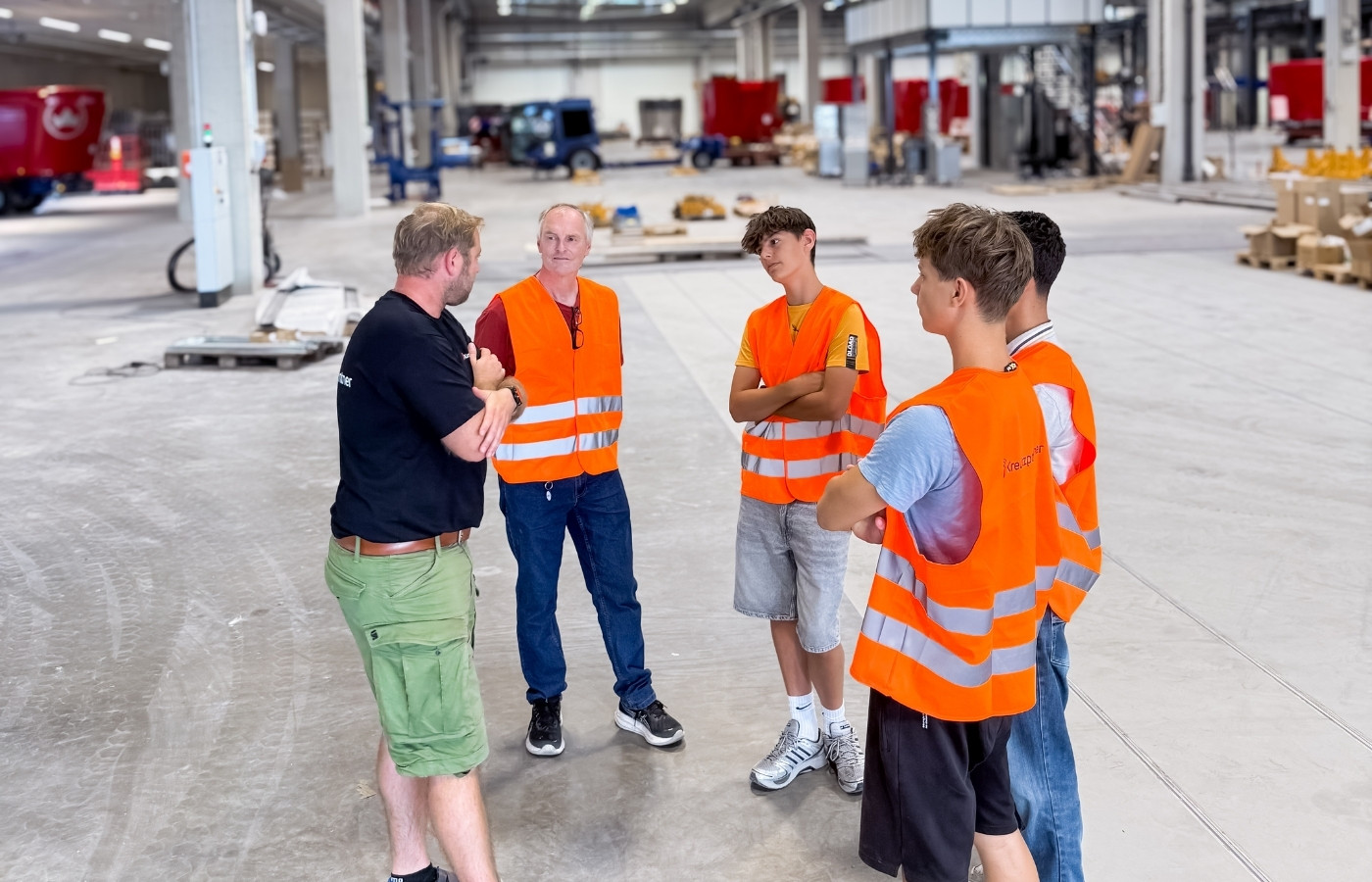 Eine Gruppe an Menschen steht im Kreis in einer großen Produktionshalle und vier davon tragen Warnwesten und hören dem anderen Herren zu.