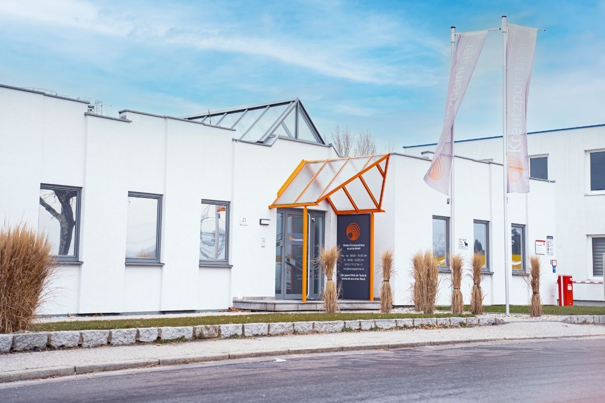 Ein Firmengebäude mit weißer Außenfassade und einem orangefarbenen Eingang mit einem Schild, auf dem das Logo und ein paar Daten zu erkennen sind.