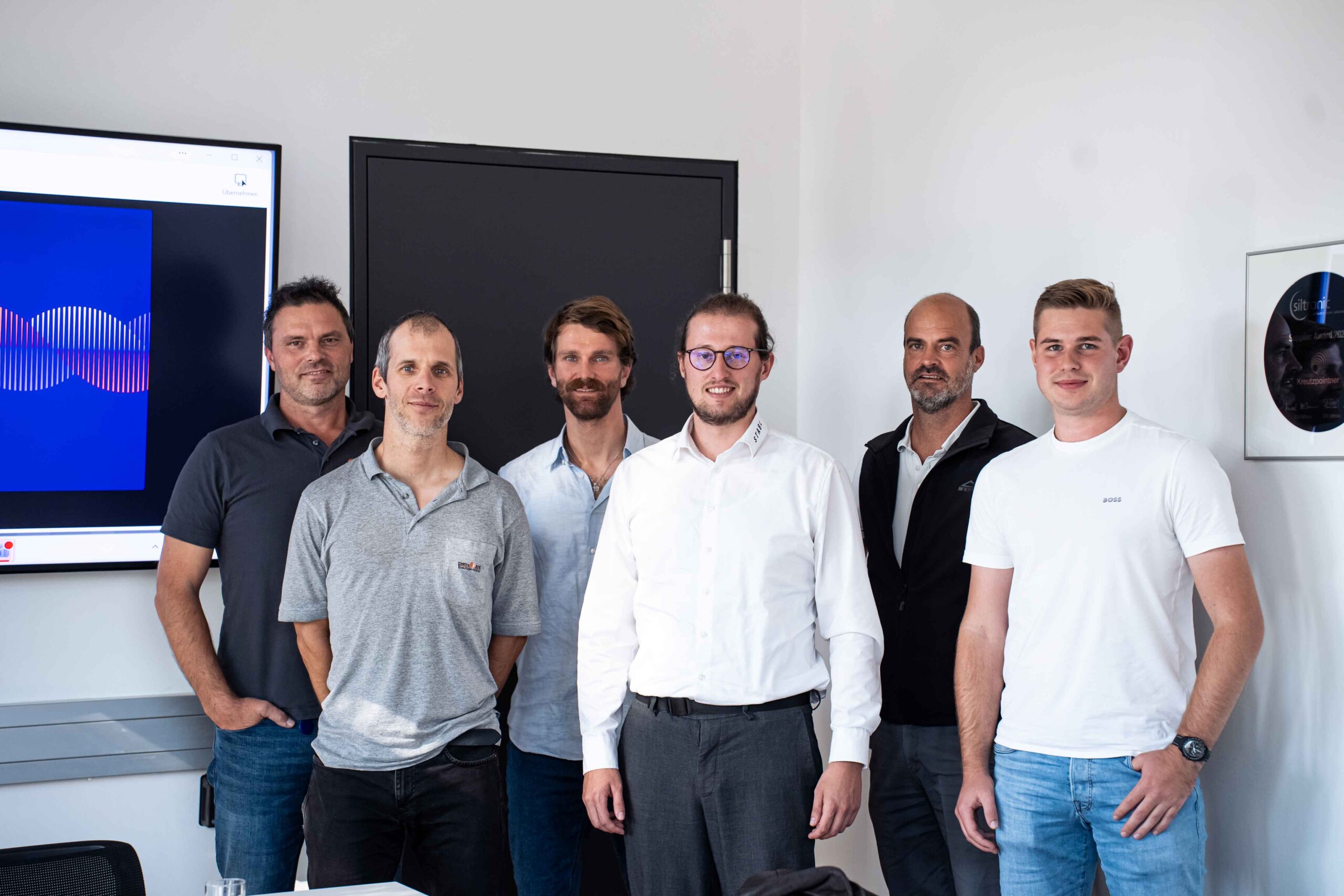 Gruppenbild von 6 Herren, die freundlich lachen und vor einem Bildschirm mit einem blauen Screen stehen.